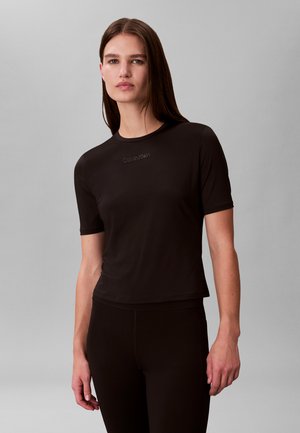 Kvinde med langt brunt hår iført en tætsiddende sort Calvin Klein kortærmet skjorte og sorte bukser, stående mod en ensfarvet grå baggrund.