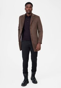 Brun ullrock över en mörk polo-tröja, kombinerad med svarta byxor och svarta snörkängor. Rocken har ett mönstrat foder.