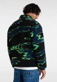 Zwarte fleecejas met een levendig groen en blauw abstract patroon, met een hoge kraag en een gestructureerd oppervlak, gezien van de achterkant.