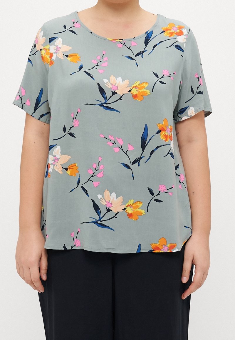 Blouse bleu clair à manches courtes avec motifs floraux orange, rose et crème, portée avec un pantalon noir, sur un fond uni.