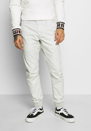 Man draagt witte joggerbroek, zwart-witte sneakers en een wit shirt met lange mouwen met zwarte en rode geborduurde manchetten.