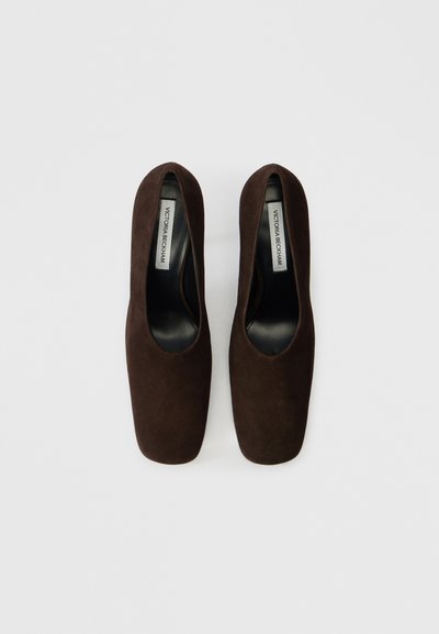 Brūni zamša pumpi ar kvadrātveida purngalu, paaugstinātu papēdi un gludu iekšējo oderi. Iezīmējas ar izgriezumu dizainu un minimālu zīmolu uz iekšējās zolītes.