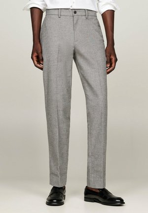 Man die lichtgrijze, op maat gemaakte broek met voorplooi en zwarte leren loafers draagt, staand tegen een neutrale achtergrond.