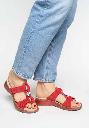 Red suede sandals with a two-strap design, featuring round metallic accents and a textured sole. Paired with light blue cuffed jeans.