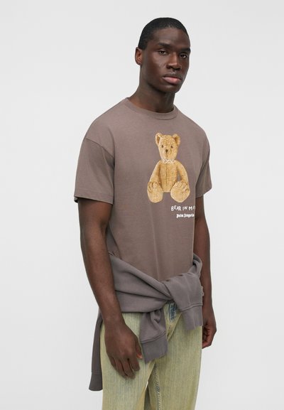 Καφέ βαμβακερό T-shirt με ένα γραφικό αρκούδας σε χρυσαφένιες αποχρώσεις και κείμενο "BEAR IN MIND", σε συνδυασμό με ανοιχτό πράσινο παντελόνι.