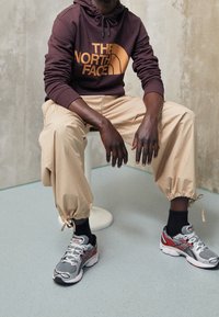 Sudadera con capucha granate de The North Face, pantalones beige entallados con puños elásticos, calcetines negros y zapatillas deportivas gris-rojas con detalle de malla.