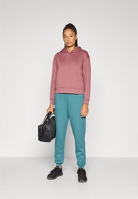 Μπορντό κοντό φούτερ με κορδόνια, πετρόλ φόρμα και μαύρα αθλητικά παπούτσια. Κρατά μια μαύρη τσάντα duffle. Ομαλή, απαλή υφή υφάσματος.