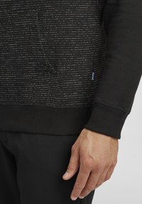 Sudadera negra con un patrón texturizado de finas líneas horizontales blancas, puños y dobladillo inferior acanalados. Etiqueta visible en el lateral.