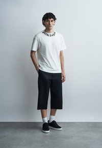 Νέος άνδρας στέκεται μπροστά σε απλή τοίχο φορώντας λευκό μπλουζάκι με γραφικό, μαύρο φαρδύ παντελόνι cropped, λευκές κάλτσες και μαύρα αθλητικά παπούτσια.