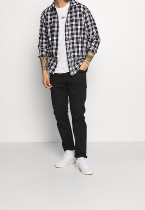 Uomo che indossa una camicia a scacchi bianca e nera, una t-shirt bianca, pantaloni neri e sneakers bianche, con tatuaggi su entrambe le avambraccia, in piedi contro uno sfondo bianco.