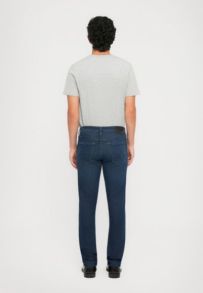 Muškarac s kovrčavom kosom nosi sivi majicu, tamnoplave traperice i crne cipele, okrenut prema jednostavnoj bijeloj pozadini.