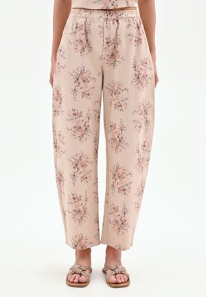 Femme portant un pantalon ample beige clair avec un imprimé floral rose et des sandales brunes tressées, debout devant un fond blanc.