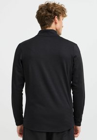 Maglietta nera a maniche lunghe con tessuto testurizzato. Presenta un colletto alto e cuciture piatte, con un orlo leggermente curvo nella parte inferiore.