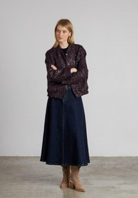 Donna in piedi con le braccia incrociate, veste una giacca di paillettes viola scuro, una lunga gonna blu scuro e stivali alla caviglia in suede beige su un pavimento di cemento.