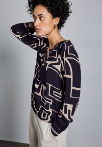 Camicia a maniche lunghe nera e beige con motivo geometrico, dotata di scollatura rotonda e polsini elasticizzati, indossata su pantaloni chiari. Design geometrico moderno.