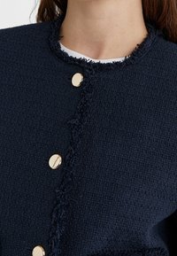 Veste texturée marine avec un bord  à franges, des boutons ronds dorés et un col ajusté. Tissu lisse avec un motif tissé.