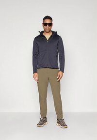 Tengerészkék cipzáras dzseki magas gallérral, zöld nadrág és túracipő. Az öltözéket napszemüveg egészíti ki. Az anyag könnyűnek és rugalmasnak tűnik.