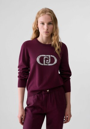 Giovane donna con lunghi capelli biondi che indossa un maglione bordeaux con logo argentato e pantaloni bordeaux abbinati, in piedi davanti a uno sfondo semplice.