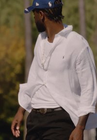 Άνδρας με λευκό πουκάμισο, μαύρο παντελόνι και μπλε καπέλο κοιτάζει προς τα πλάγια έξω με θολά δέντρα στο φόντο.