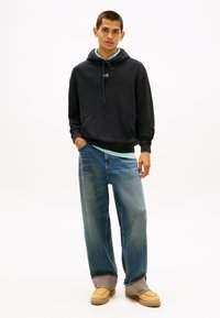 Felpa con cappuccio nera con coulisse, tasca frontale e camicia a strati blu-verde sotto. Jeans blu larghi e scarpe beige senza lacci completano il look.