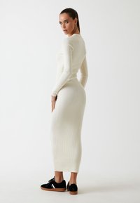 Geribbelde, langwerpige midi-jurk in off-white, gecombineerd met zwarte sneakers met een witte streep en een bruine zool. Figuratieve pasvorm.