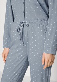 Ljusblå pyjamasset med vita prickar, bestående av en skjorta med knappar och byxor med resårmidja, satinband och fickor.