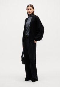 Masiven črn pleteni kardigan preko sive rolke, v kombinaciji z ohlapnimi črnimi hlačami. Model drži črno torbico čez ramo. Minimalističen stil.