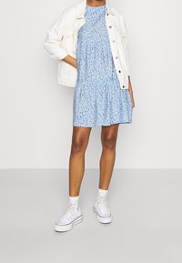 Blommig blå klänning med volangkant, kombinerad med en vit denimjacka. Vita högklackade sneakers och ankelsockor avslutar outfiten.