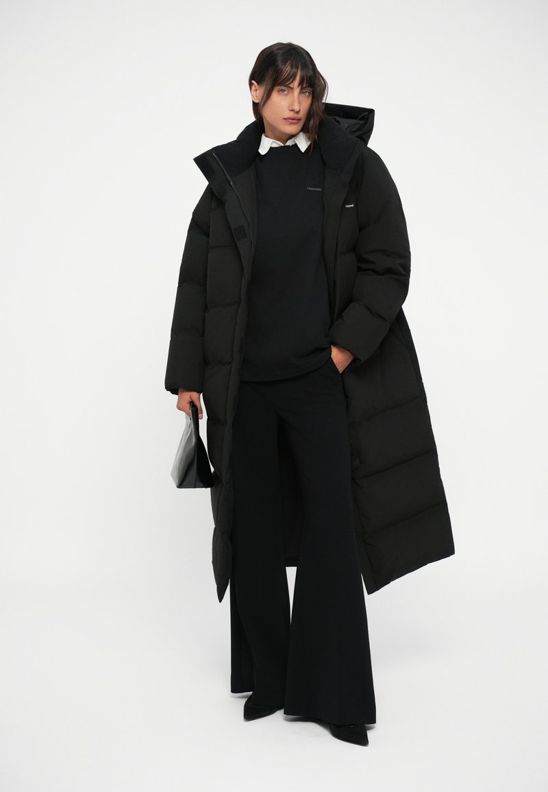 Black puffer coat with a high collar, oversized design, textured fabric; paired with a black sweater and wide-leg trousers. Black bag.