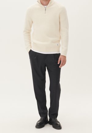 Homme portant un pull à col zippé côtelé crème, un sous-vêtement blanc, un pantalon à plis foncé, et des mocassins en cuir noir, debout les mains dans les poches.