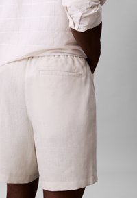 Mann trägt weiße Leinenhose mit einer Gesäßtasche und ein passendes weißes Hemd mit hochgekrempelten Ärmeln vor einfachem Hintergrund.