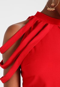 Top rosso senza maniche con ritagli sulle spalle e spalline che ricadono a cascata. Tessuto liscio con collo alto e dettagli di design eleganti.