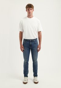 Kék farmernadrág egyenes szabással és öt zsebbeli, sima fehér pamut pólóval és fehér sportcipővel párosítva.