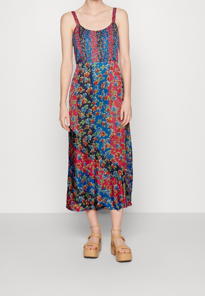 Femme portant une robe sans manches, longue jusqu'aux chevilles, à motif floral avec des panneaux verticaux multicolores bleus, rouges et noirs, et des sandales compensées beiges.