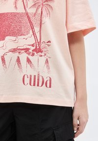 Ανοιχτό ροζ πουκάμισο με κόκκινο σχέδιο φοίνικες, παραθαλάσσια σκηνή και το κείμενο "VANA cuba", φορεμένο με μαύρο παντελόνι, χέρι ξεκουρασμένο στο πλάι.