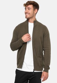 Olivengrøn lynlås cardigan med zigzag tekstur, ribbede manchetter og kant, båret over en hvid skjorte, lavet af et glat, let materiale.