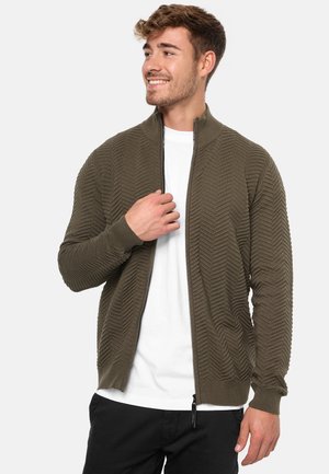 Cárdigan verde oliva con cremallera y textura en zigzag, con puños y dobladillo acanalados, llevado sobre una camisa blanca, fabricado en un material suave y ligero.