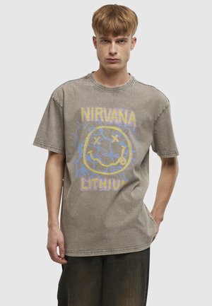 Mladý muž nosi vyblednuté sivé tričko Nirvana "Lithium" s žltým usmievavým smajlíkom a čierne nohavice, stojaci na jednoduchom pozadí.