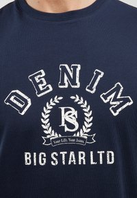 Tamsiai mėlynas medvilninis marškinėliai su grafiniu dizainu, kuriame balti užrašai "DENIM", "BS" ir "BIG STAR LTD", apsupti laurų rašto.