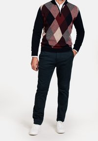 Męski sweter dzianinowy z kołnierzem na zamek, w wzór argyle w kolorach bordowym, szarym i granatowym, zestawiony z ciemnozielonymi spodniami i białymi trampkami.