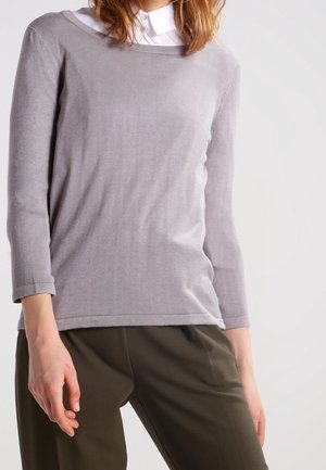 Kobieta ubrana w jasnoszary sweter z długim rękawem na białą koszulę z kołnierzykiem oraz oliwkowozielone spodnie, stoi na jednolitym tle.