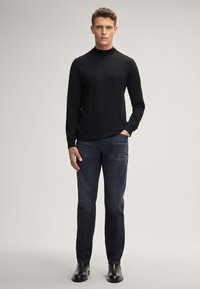 Pull en col roulé noir en tissu lisse, associé à un jean bleu foncé et des chaussures en cuir noires. Design simple, ajusté, avec un minimum de détails.