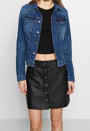 Kvinde iført blå denimjakke over sort crop top og sort knappet lædernederdel, stående mod en ensartet lys baggrund.