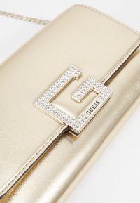 Pochette métallique dorée à finition brillante, ornée d'un logo "G" proéminent décoré de strass, avec une fine chaîne pour la porter.
