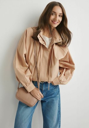 Młoda kobieta z długimi brązowymi włosami, ubrana w beżową kurtkę i niebieskie dżinsy, nosząca małą beżową torebkę na ramię, uśmiechająca się na tle jednolitej ściany.