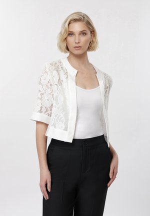 Kvinde med kort blondt hår iført hvid blondecardigan over hvidt tanktop og sorte bukser mod en ensfarvet hvid baggrund.