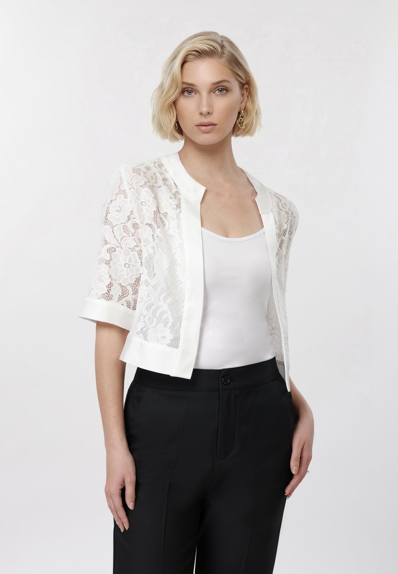 Femme aux cheveux blonds courts portant un cardigan en dentelle blanche par-dessus un débardeur blanc et un pantalon noir sur un fond blanc uni.