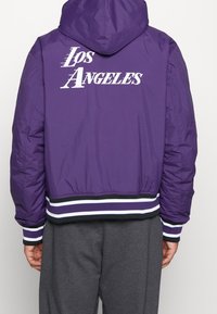 Lila puffjacka med huva, med "Los Angeles" i vit text på baksidan, samt randiga ribbade ärmar och nederkant i svart och vitt.