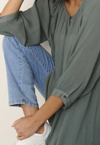 Grüne plissierte Bluse mit Schlüssellochausschnitt, lockerer Passform und dreiviertel Ärmel, kombiniert mit hellblauen Jeans-Ankle-Pants und weißen Sneakers.