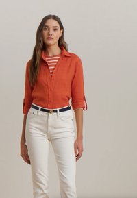 Orange button-up shirt with rolled sleeves, layered over a red and white striped top. Paired with white jeans and a navy belt with gold hardware.